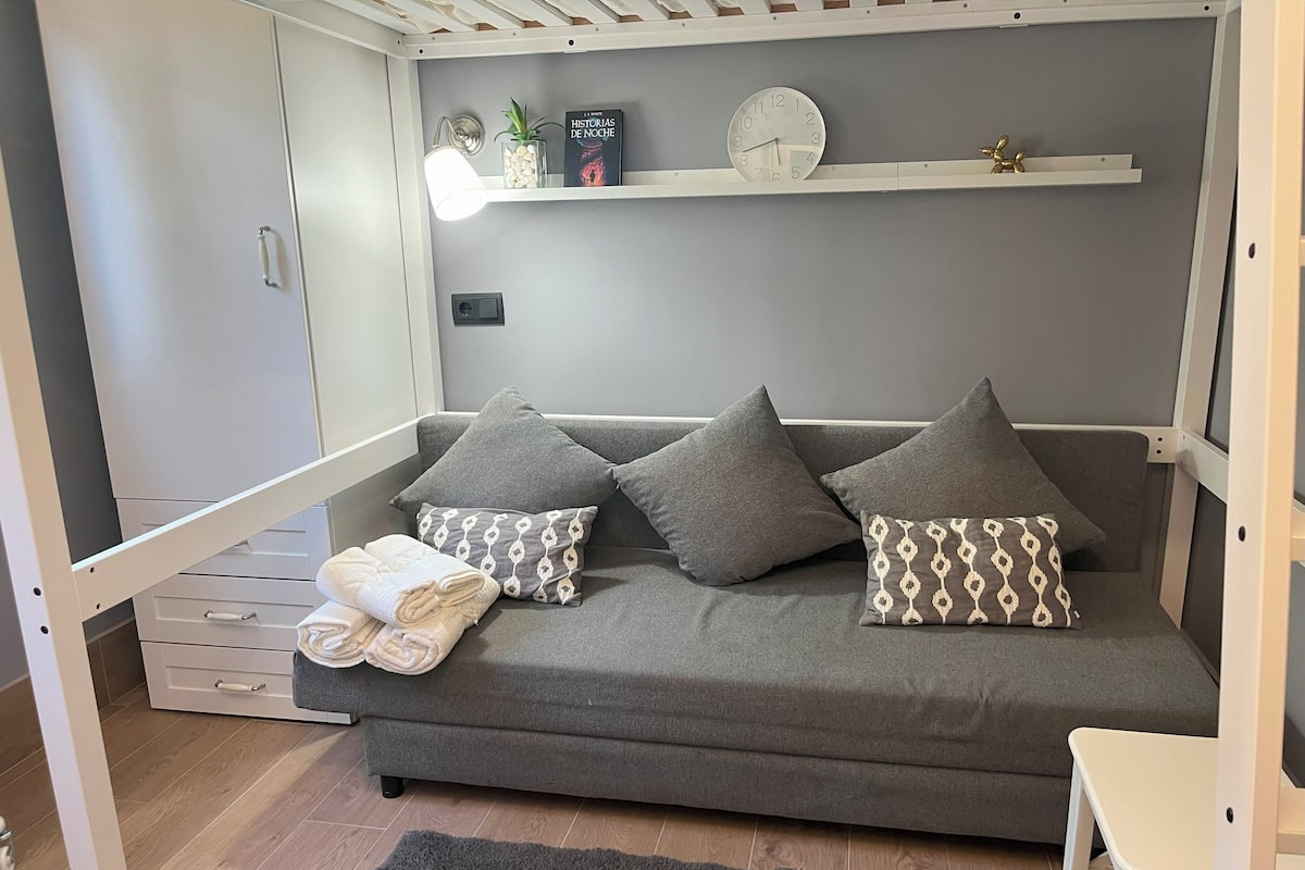 A modern living room with a grey sofa, patterned cushions, a small white table, and a shelf with decorative items.