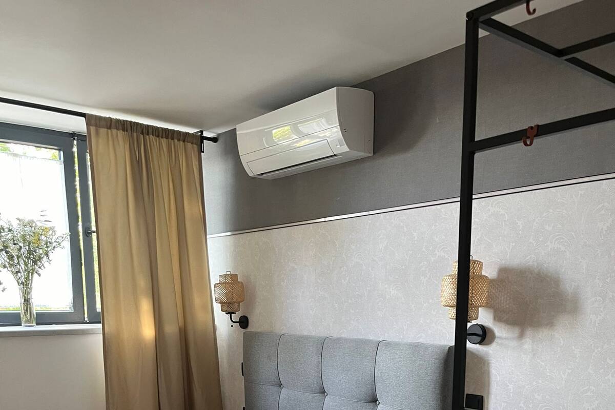 A compact room with a bed, a window with curtains, a wall-mounted air conditioner, a small sink, and a yellow basket.