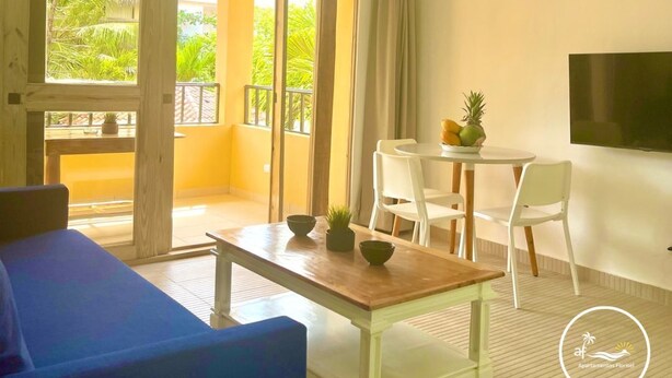 A room with a wooden ceiling, a ceiling fan, a TV mounted on the wall, a dining table with chairs, and a sliding glass door leading to an outdoor area with plants. - A3 Modern apt on the Beach in Punta Cana (Punta Cana)