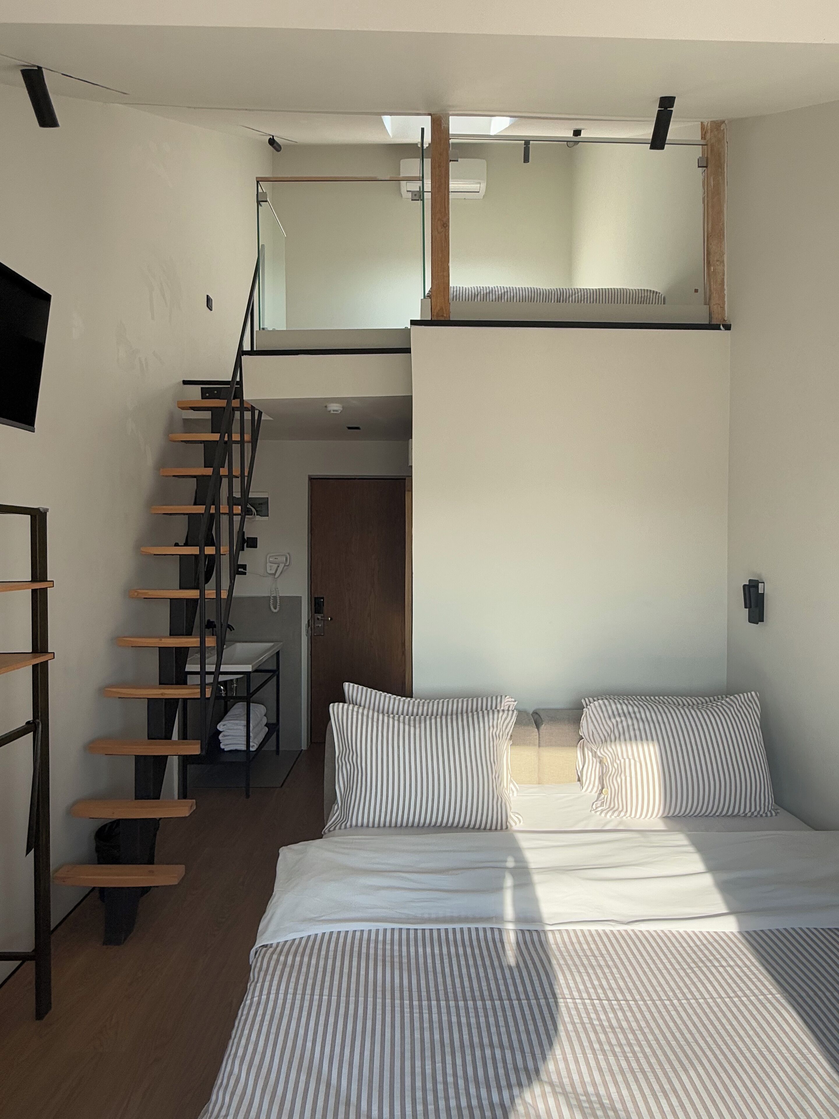 A modern bedroom with a loft-style bed, a wooden ladder, and a small bathroom.
