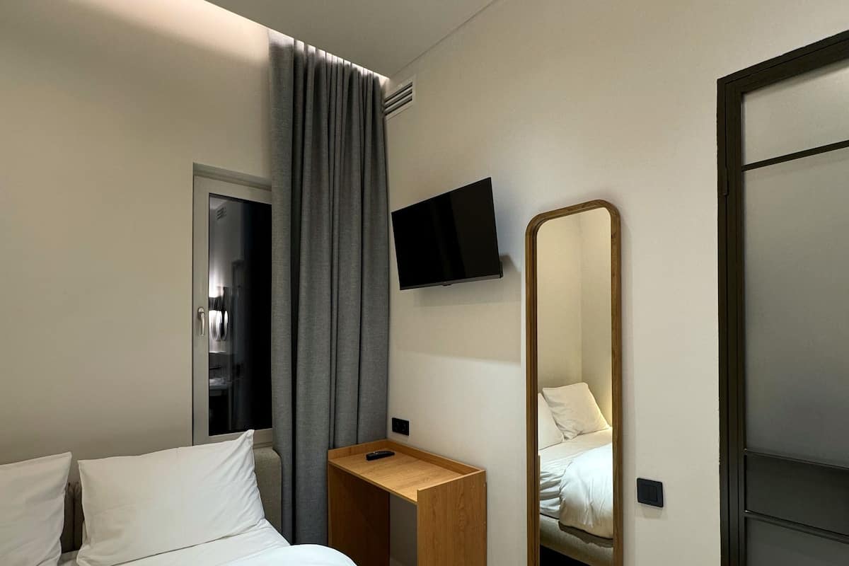 A modern hotel room with a bed, a wooden nightstand, a flat-screen TV, a mirror, and a door with a curtain.