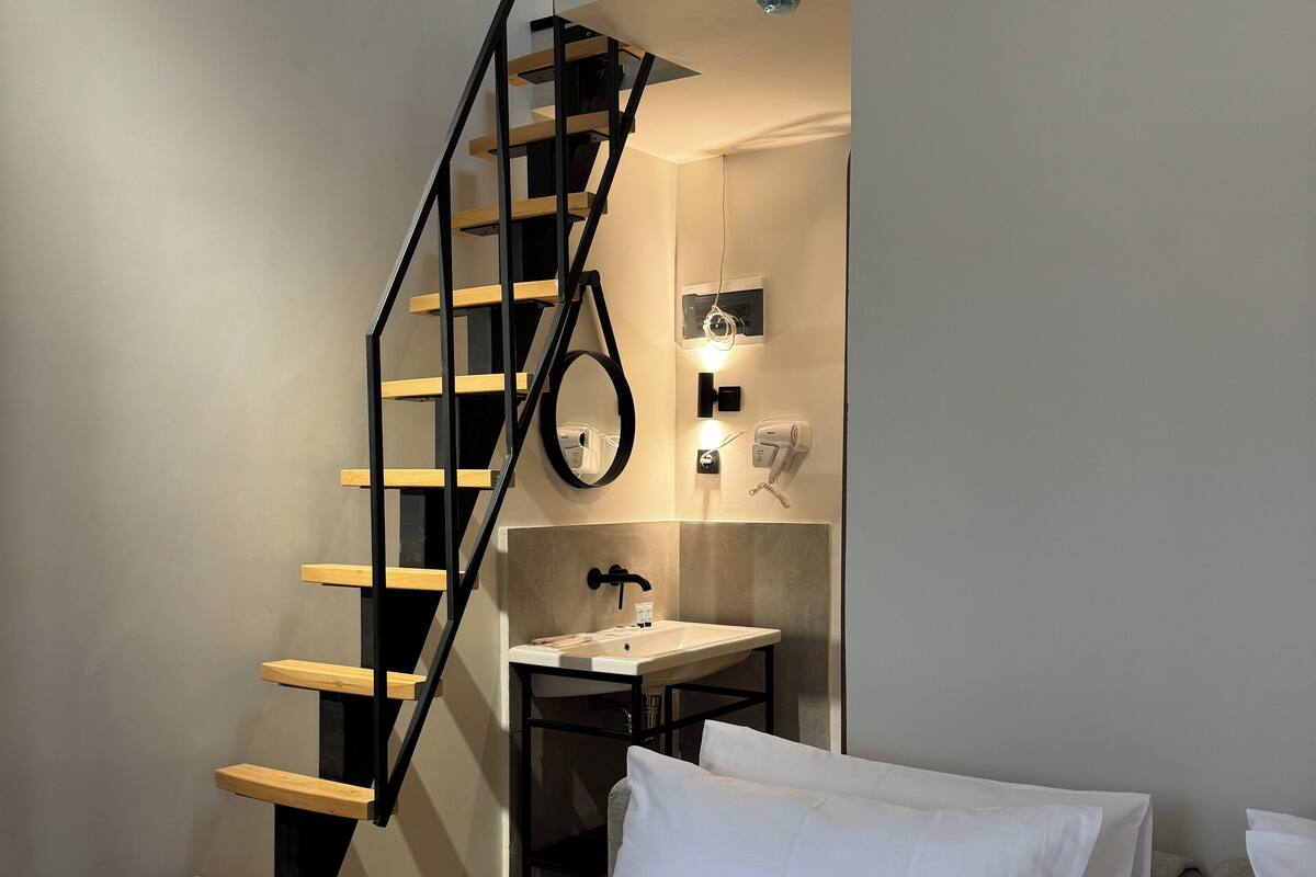 A bedroom with a bed, white pillows, a wooden staircase, a small bathroom with a sink and a mirror.