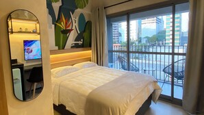 A hotel room with a bed, a desk with a computer, a TV, a large window with a view of the city, and a ceiling fan.