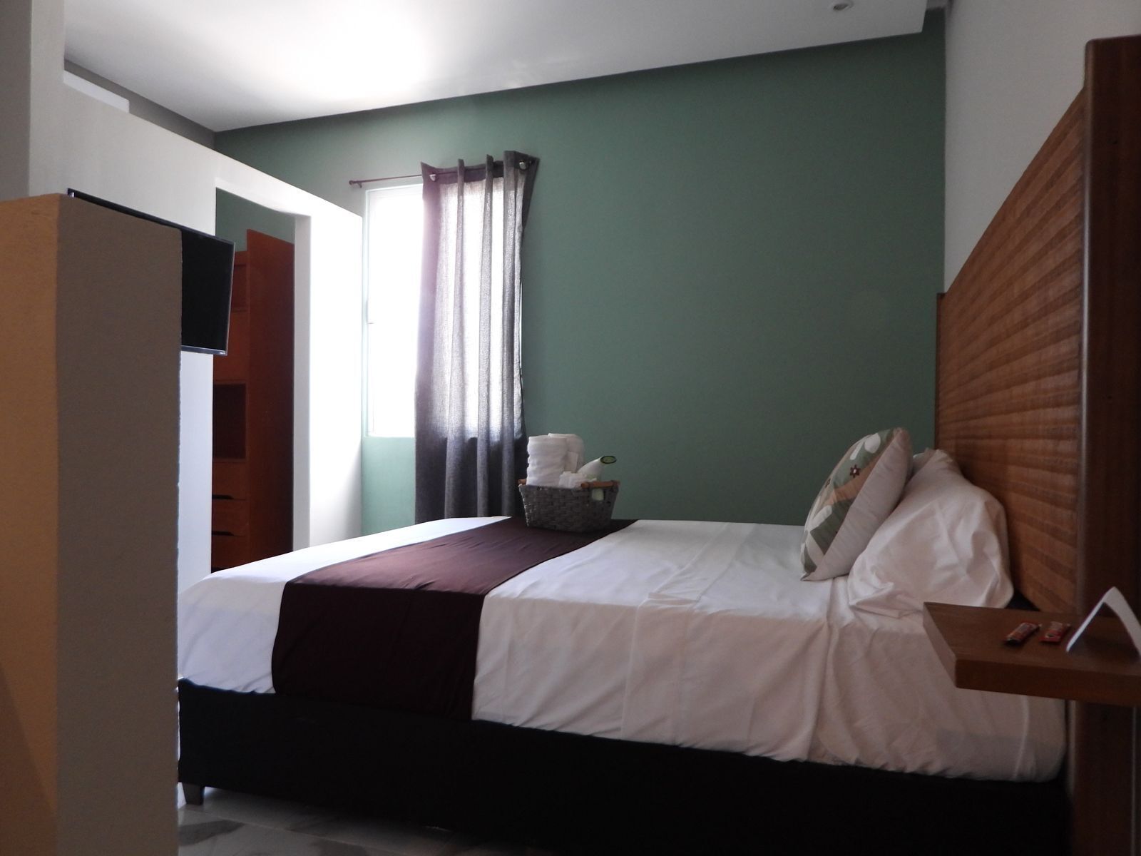 A bedroom with a bed, a wooden headboard, a bedside table, a basket with towels, and a window with curtains.