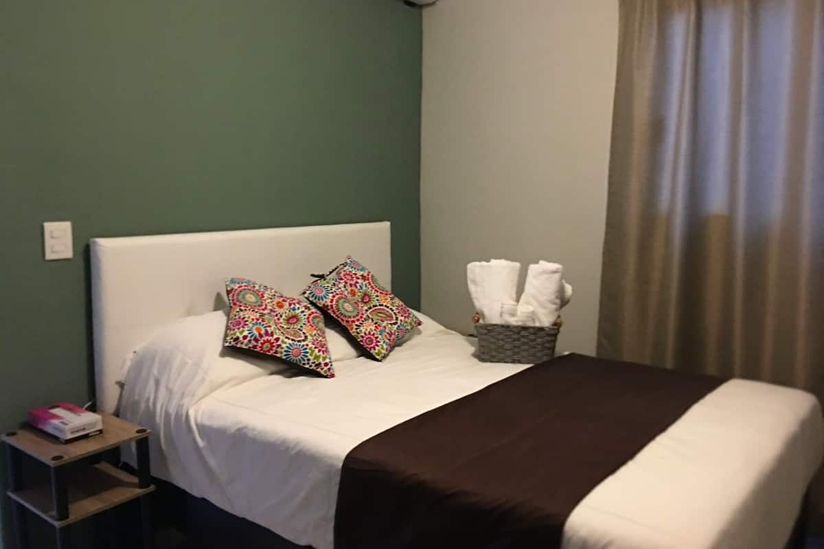 Un lit bien fait, avec une tête de lit blanche, deux coussins colorés, un plaid marron et un panier rempli de serviettes pliées, posé sur une table de chevet.