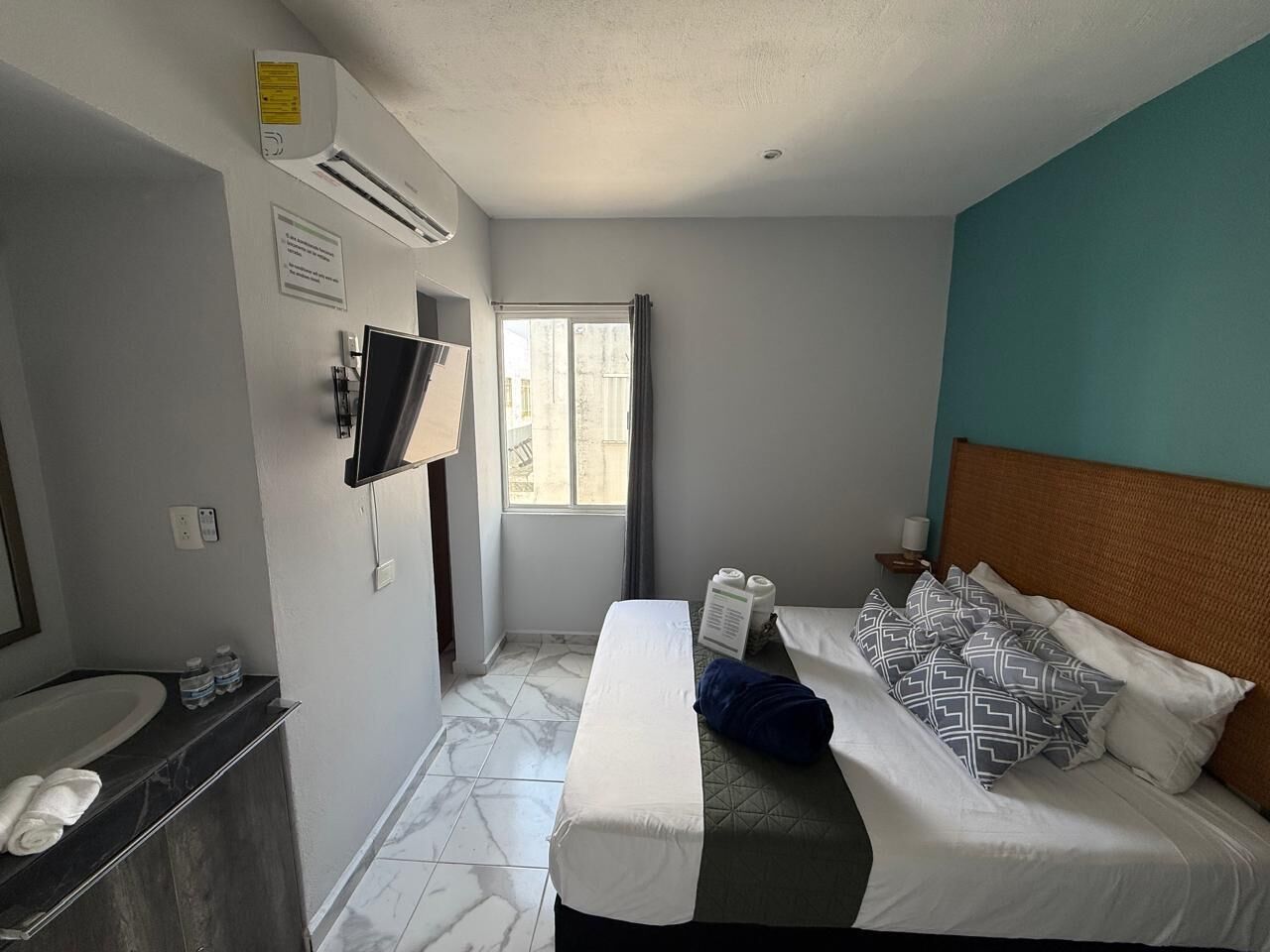 A hotel room with a bed, a TV mounted on the wall, a bathroom with a sink and mirror, and a window with curtains.