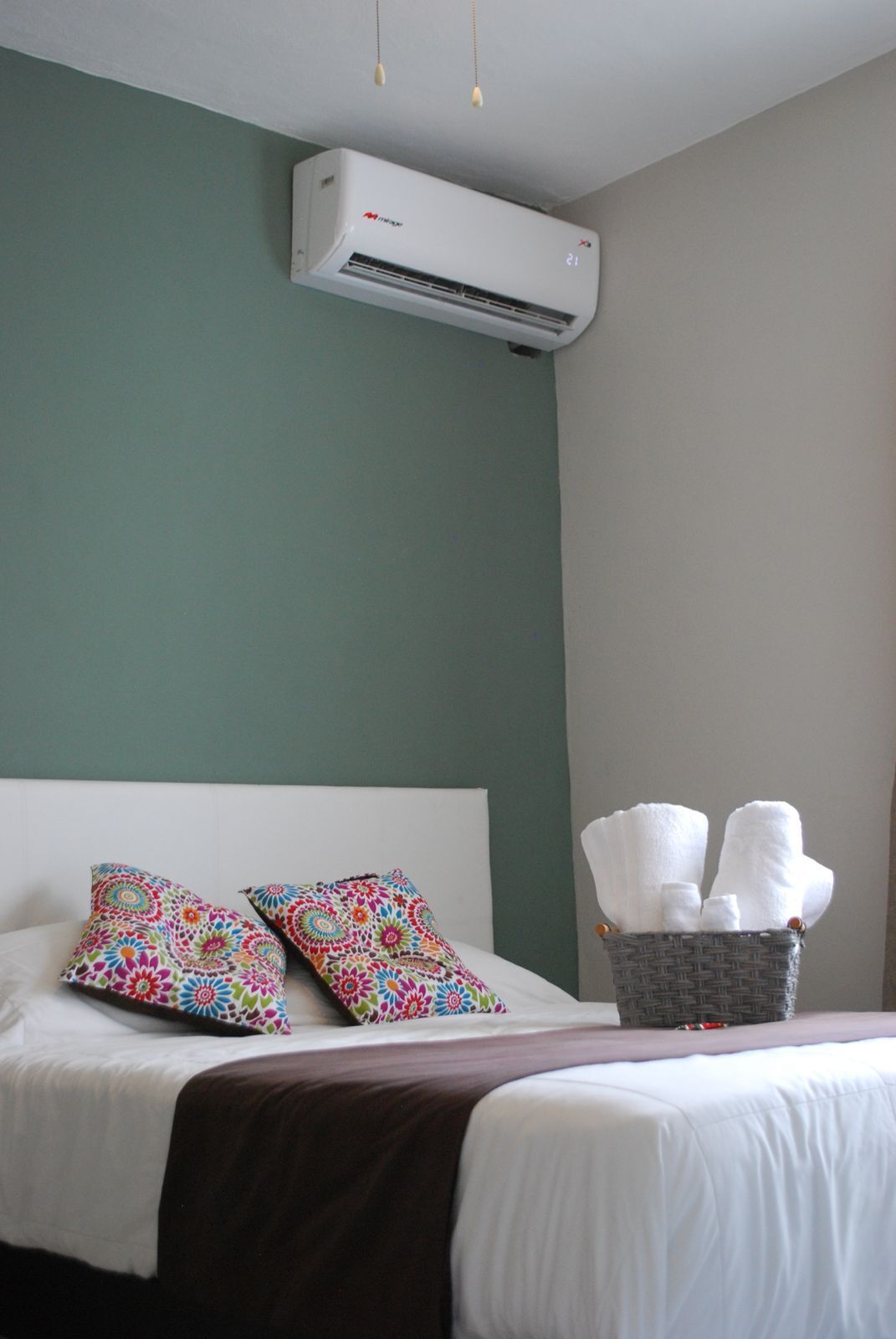 A hotel room with a bed, two colorful pillows, a basket of towels, and an air conditioning unit on the wall.