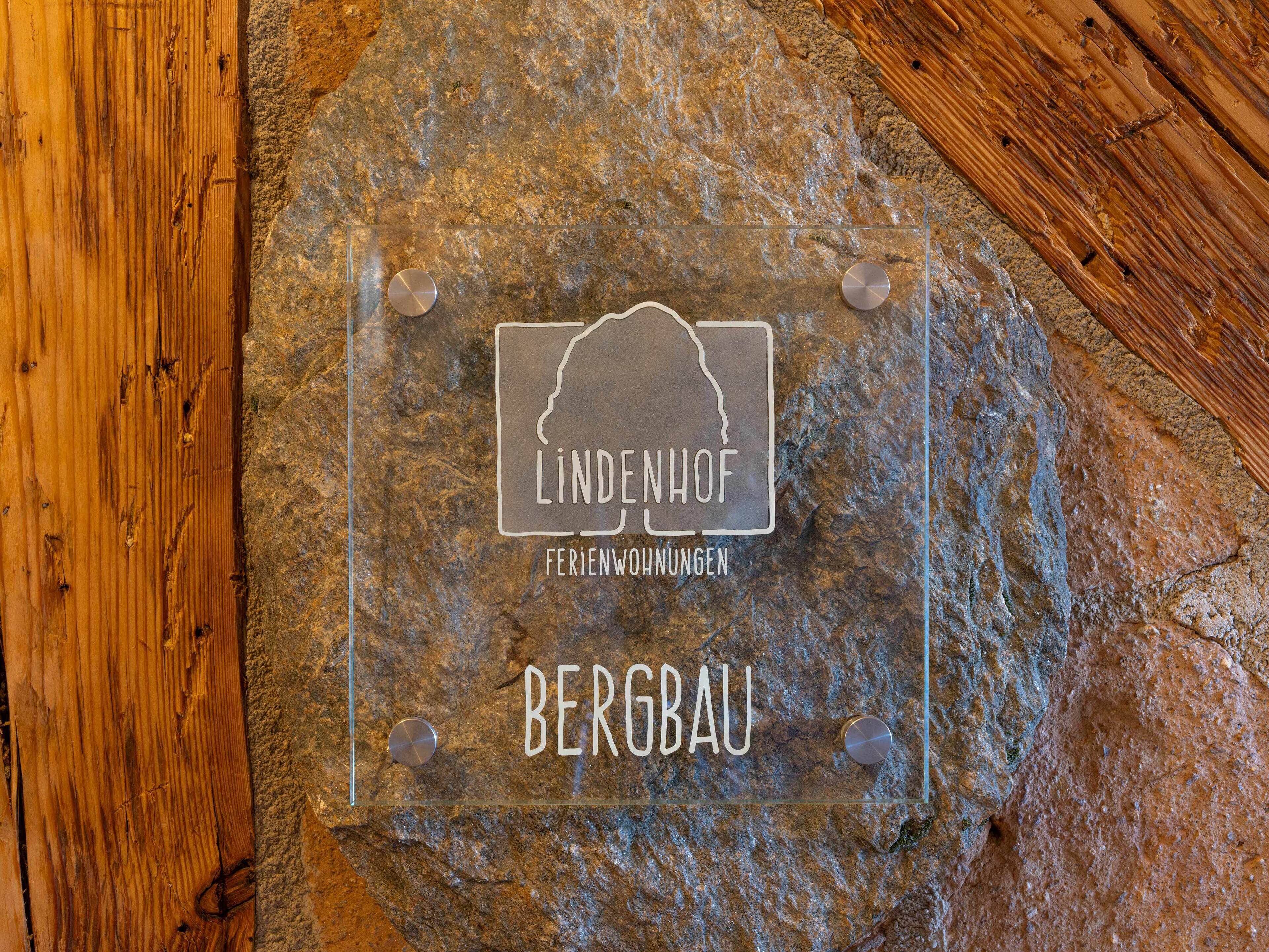 Een transparante glazen plaat met de tekst “LINDENHOF FERIENWOHNUNGEN BERGBAU” vastgemaakt aan een stenen muur.