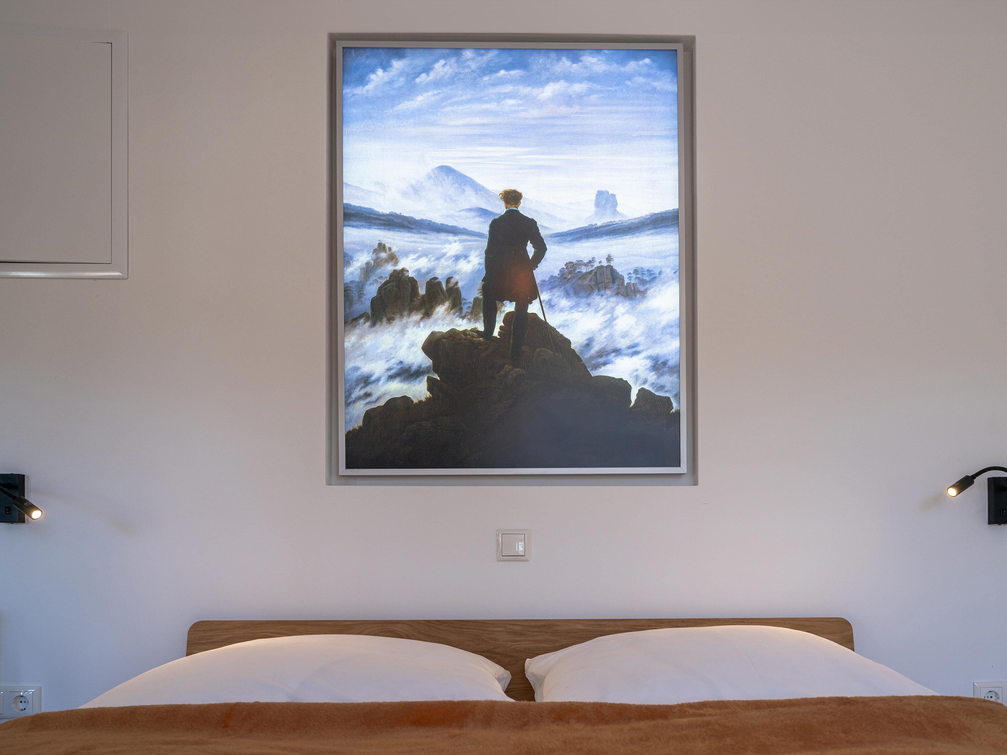 Een slaapkamer met een groot bed, een lijst met een schilderij van een man op een klif, en een nachtlampje aan de muur.