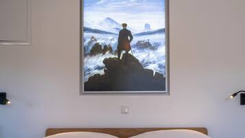 Een slaapkamer met een groot bed, een lijst met een schilderij van een man op een klif, en een nachtlampje aan de muur.