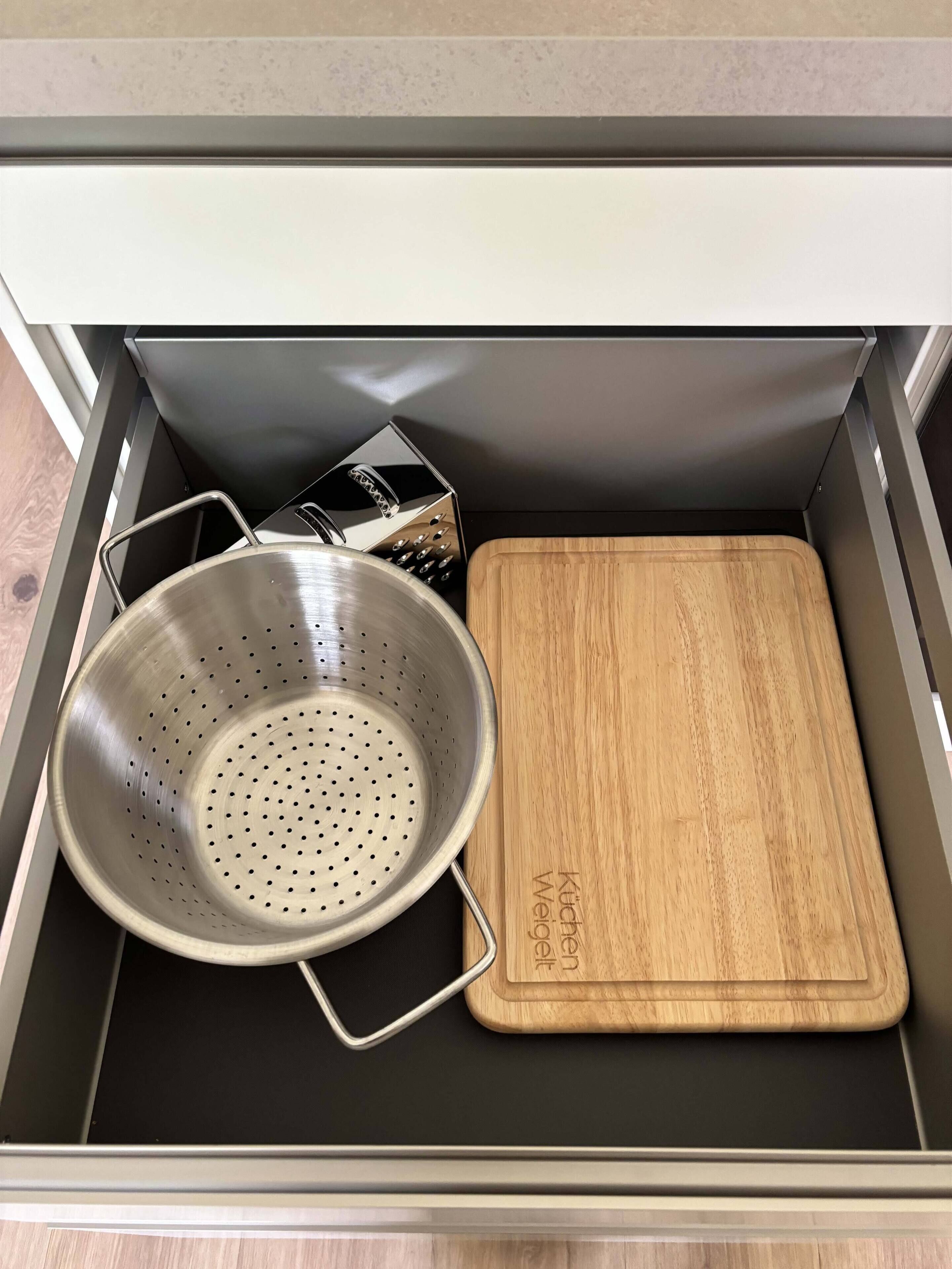A kitchen drawer with a colander, a wooden cutting board, and a grater.