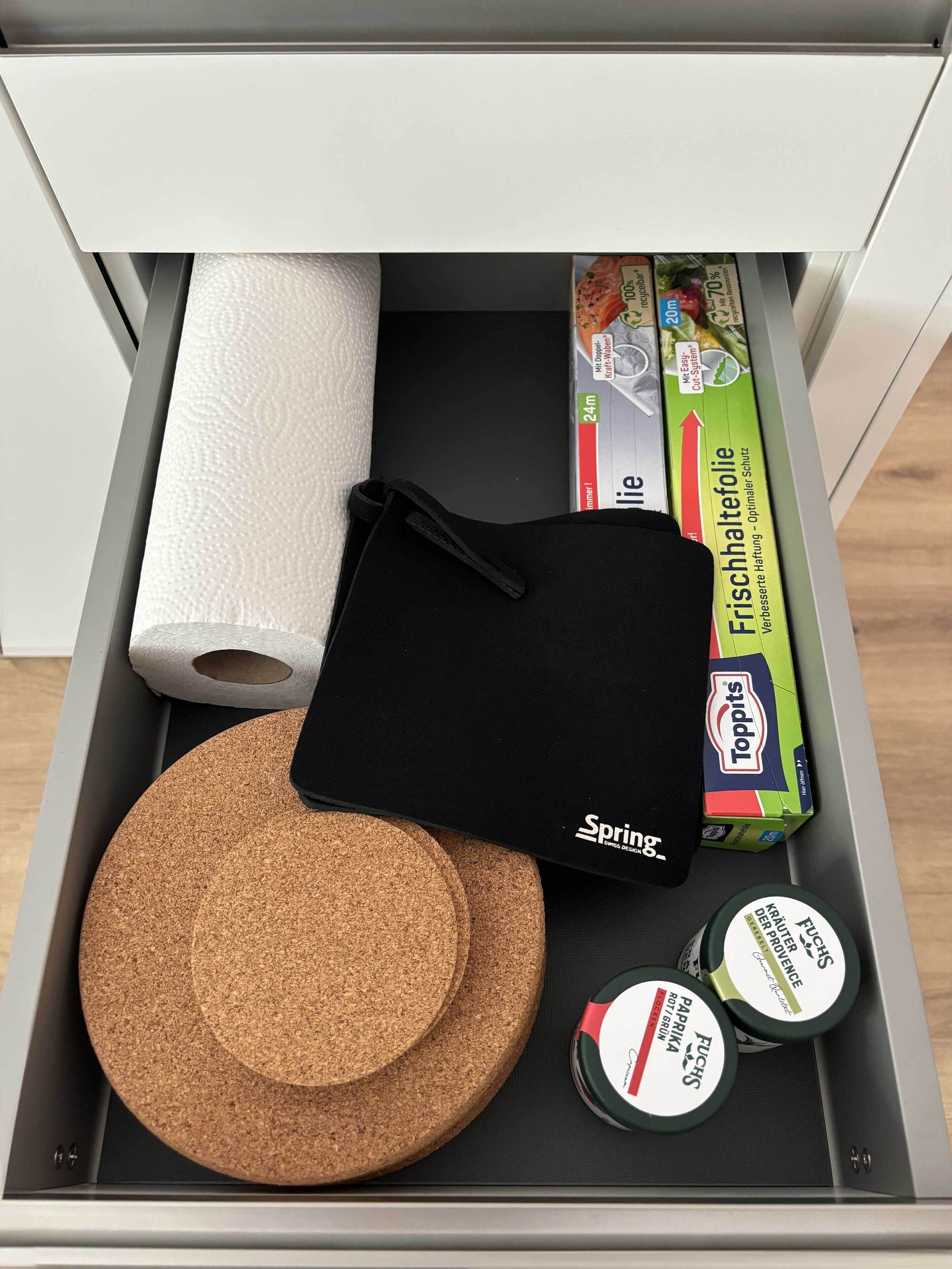 A drawer containing a black bag, two cork coasters, a roll of paper towels, a box of Toppits, and two containers of adhesive.