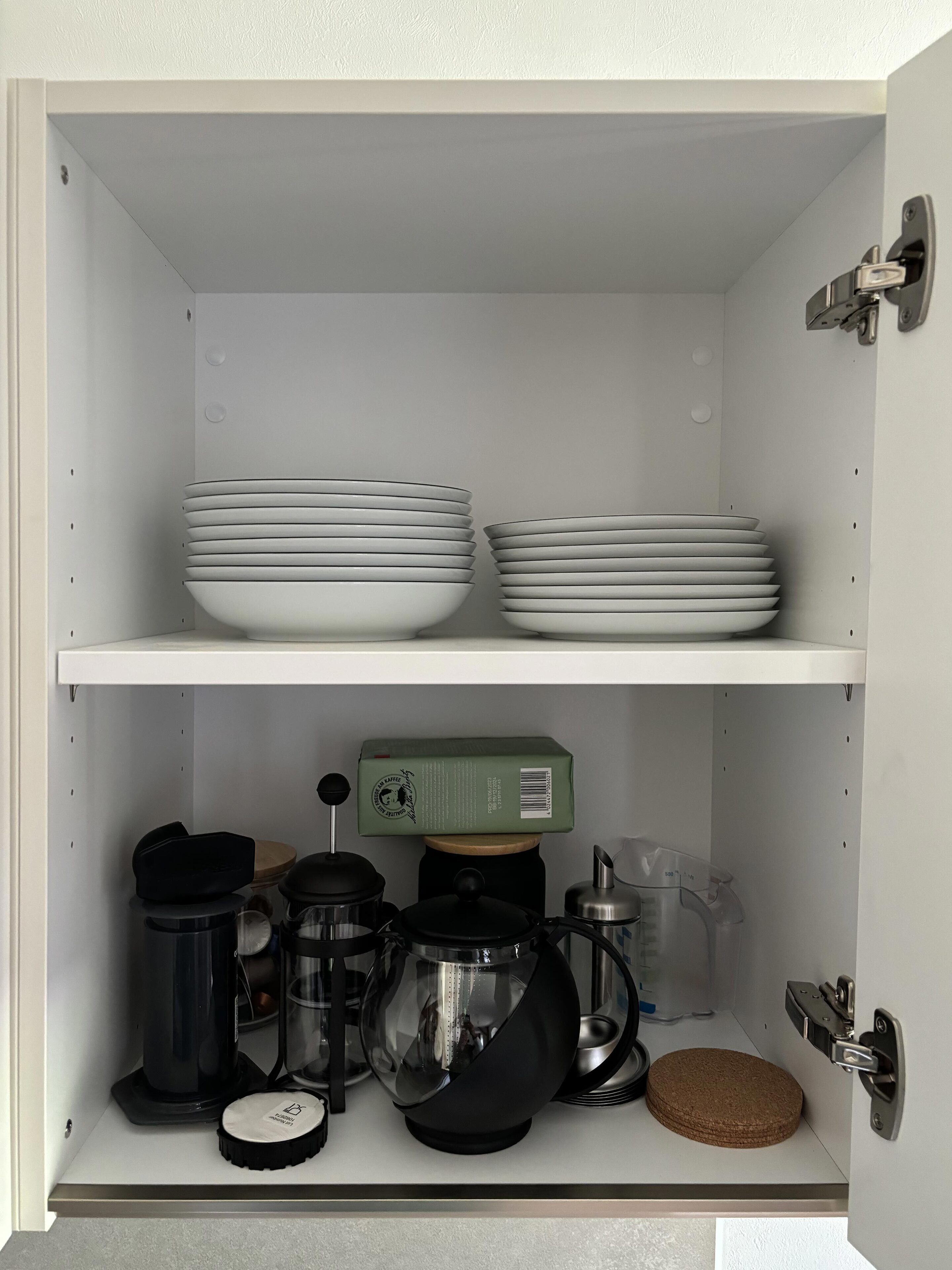 Ein Küchenschrank mit weißen Regalen, der gestapelte weiße Teller, eine grüne Kiste, eine Kaffeemaschine, einen Wasserkocher, einen Mixer und eine Korkmatte enthält.