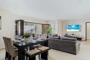 A modern living room with a dining area, a grey sofa, a flat-screen TV, and a large mirror. - Private Pool Oasis! Secluded 4BR w Patio & Parking (Santa Cruz)