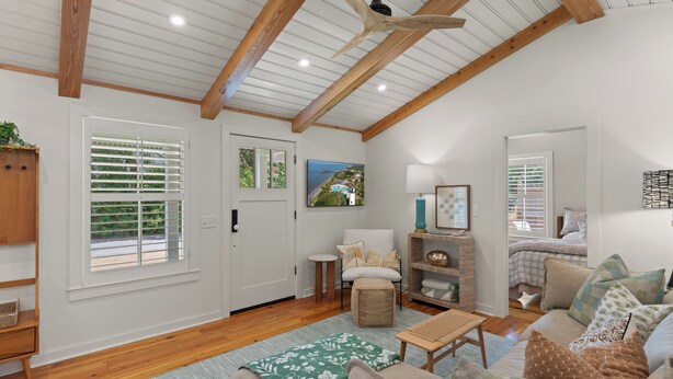 A spacious living room with a high ceiling, wooden beams, a ceiling fan, a large window with blinds, a comfortable sofa, a small table, and a cozy reading chair.