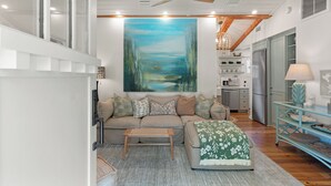A modern living room with a beige sofa, a wooden coffee table, and a green patterned throw. A large painting of a landscape is on the wall, and there is a kitchen area in the background.