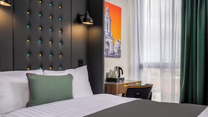 A hotel room with a large bed, a wall-mounted light fixture, a painting, a desk with a kettle, and a view through sheer curtains. - Point A Dublin The Liberties (Dublin)