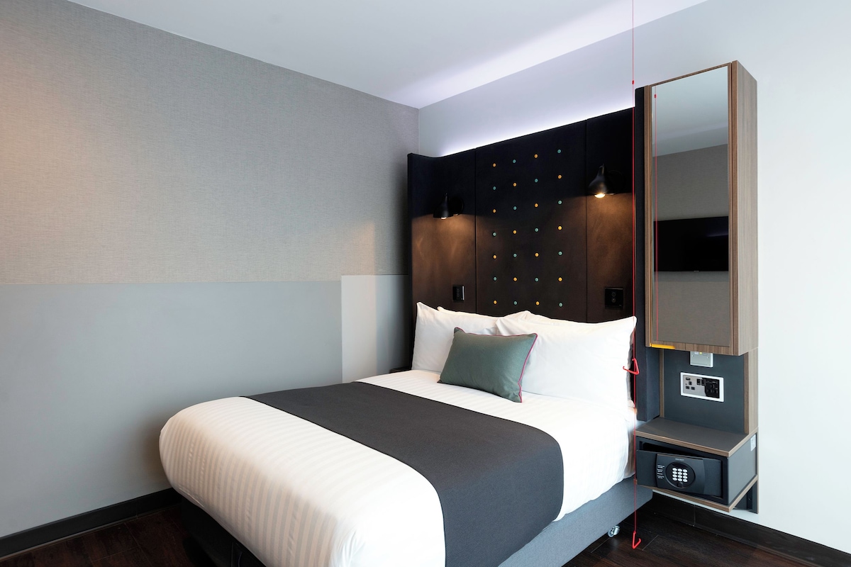 A modern hotel room with a large bed, a nightstand with a lamp, a mirror, and a built-in safe.