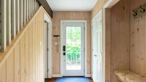 Un pasillo interior de madera con una puerta que da al exterior, un tapete azul estampado y un banco junto a la puerta.