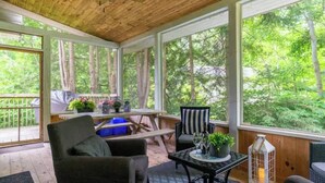 Un área de estar al aire libre techada, con mobiliario de madera, ventanas amplias y vistas al bosque.
