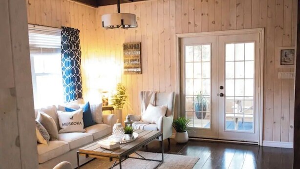 Un salón acogedor con sofá, sillón y mesa de centro. Se observa una puerta con paneles de vidrio y un letrero en la pared.
