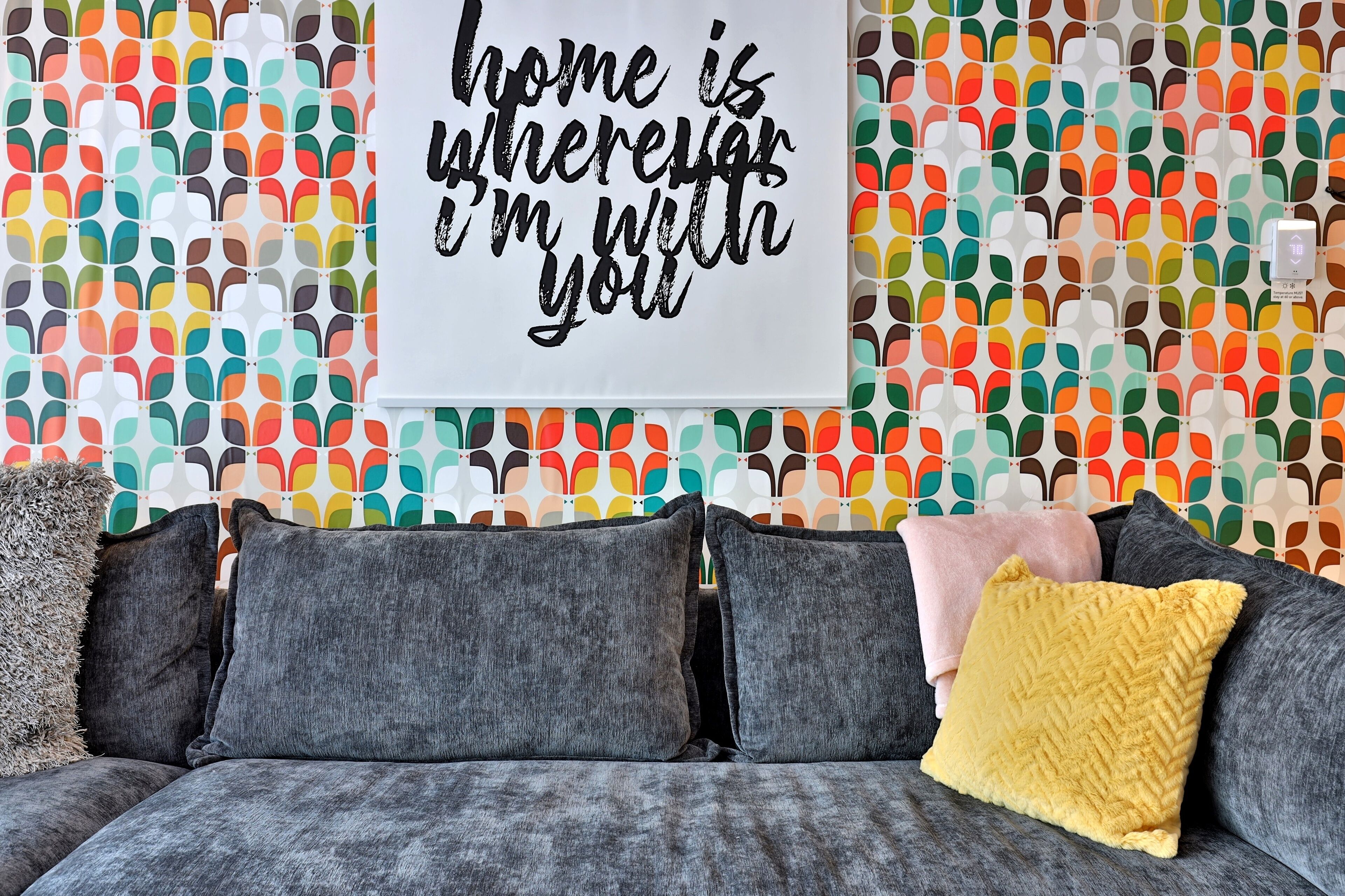 A living room with a colorful abstract wall pattern, a gray sofa with multiple cushions, and a motivational quote on the wall.