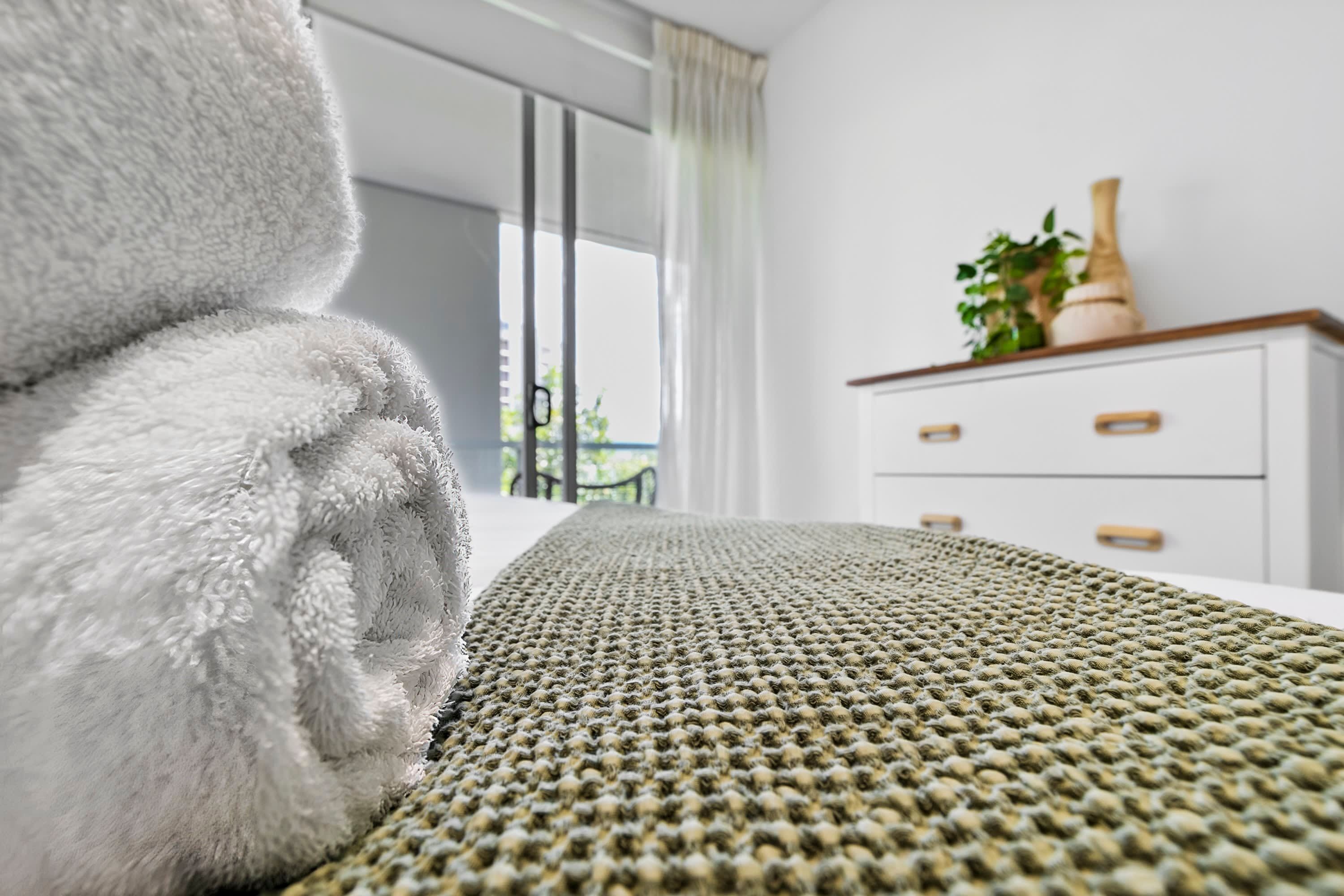 Una cama bien hecha con un edredón texturizado, un aparador blanco con cajones y una planta en maceta encima.