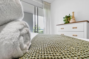 Een net opgemaakt bed met een gebreide deken, een witte ladekast en een potplant erop.