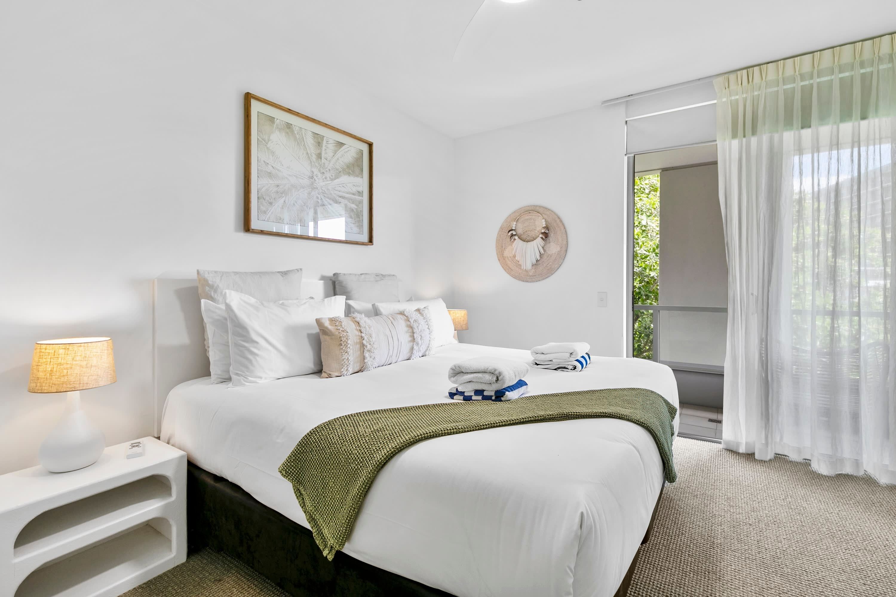 Habitación de hotel con una cama bien hecha, una lámpara de noche, una obra de arte enmarcada y vistas a zonas verdes a través de la ventana.