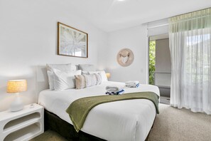Een hotelkamer met een netjes opgemaakt bed, een bedlampje, een kunstwerk in een lijst en uitzicht op groen door het raam.
