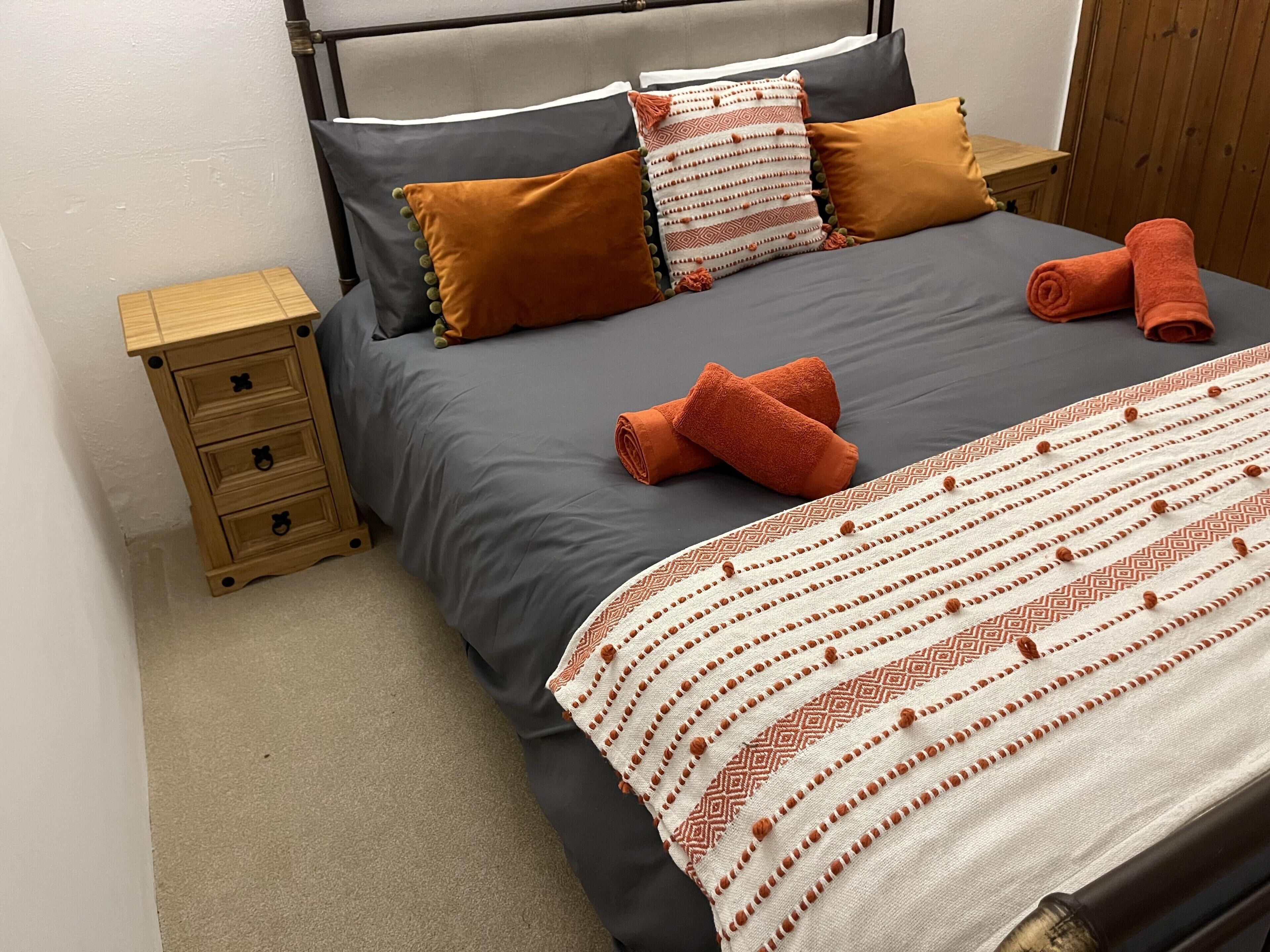 Een net opgemaakt bed met een grijze en oranje deken, twee kussens en twee opgerolde handdoeken.