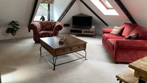 Una sala de estar estilo loft acogedora, con techo alto, un ventanal grande, un televisor, un sofá rojo, un sillón estampado y una mesa de centro de madera.