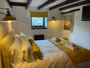 Een slaapkamer met een bed, kussens, een nachtkastje met handdoeken, een raam en een schilderij aan de muur.