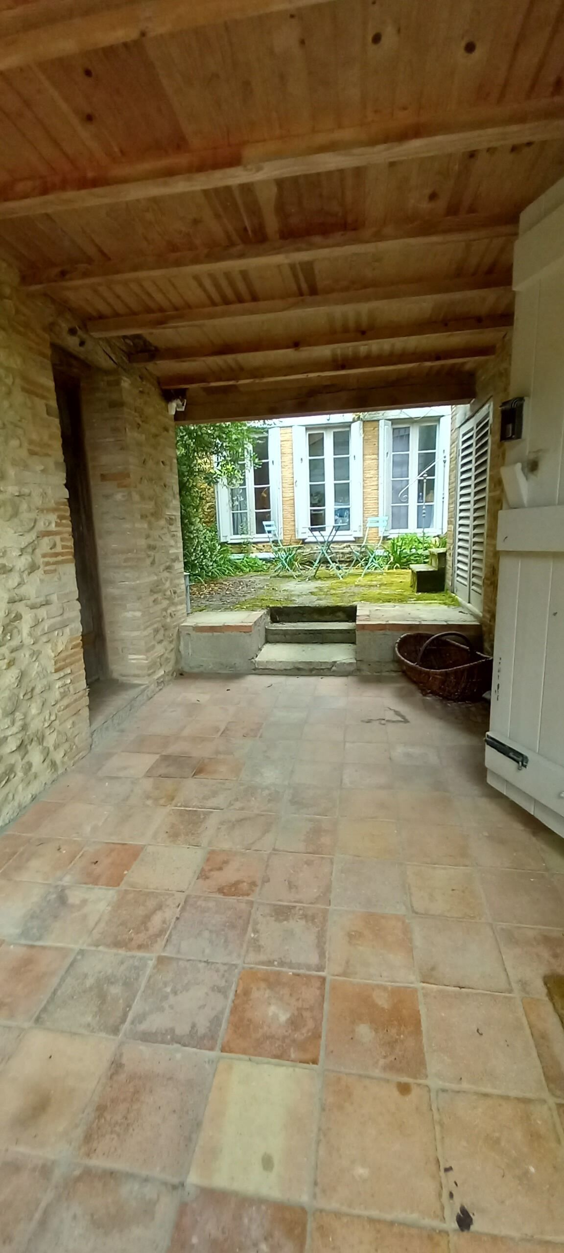 Eine offene Terrasse mit Steinboden, Holzbalken und Blick auf ein Haus mit Garten.