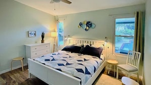 Una camera da letto con un letto bianco, un comodino, una sedia, una finestra e una ventola a soffitto.