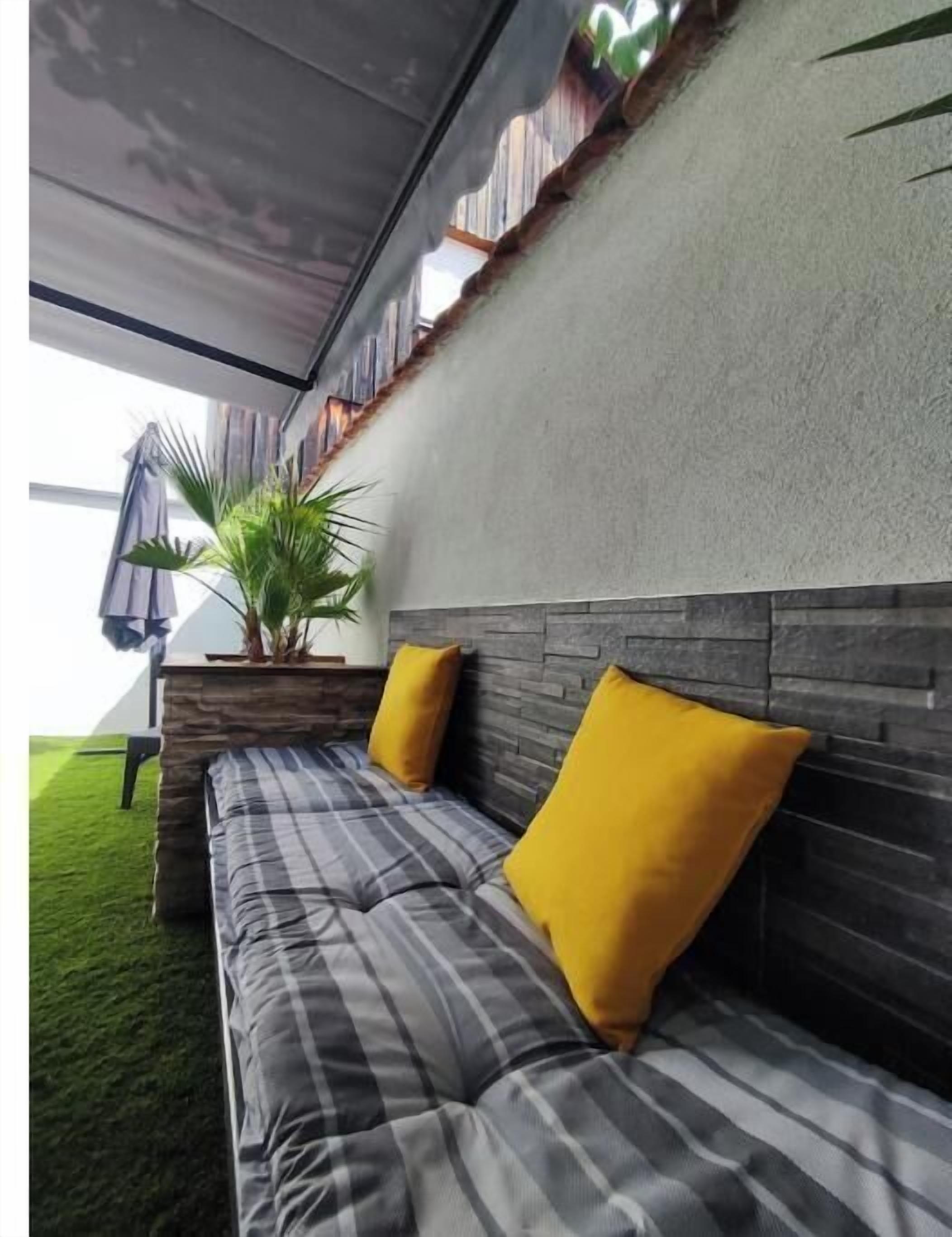 A modern outdoor seating area with a grey and black striped sofa, two yellow cushions, a potted plant, and a grey awning.