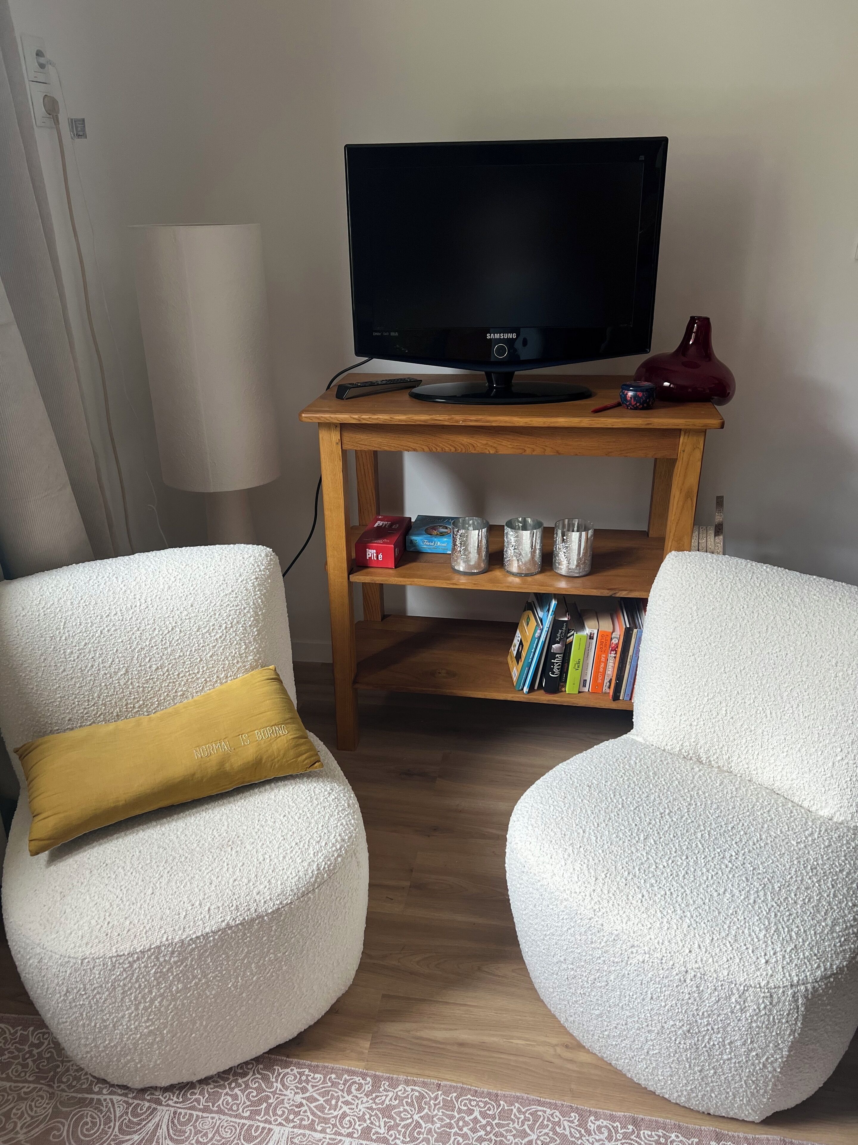 A living room with a television on a wooden stand, two white armchairs, a small table with a red vase, and a bookshelf with books.