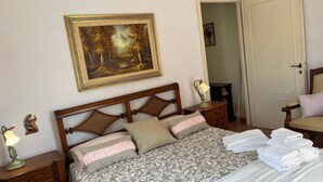 A neatly made bed with a patterned bedspread, a nightstand with a lamp, a framed painting on the wall, and a door in the background. - Casa Nettuno (Nettuno)