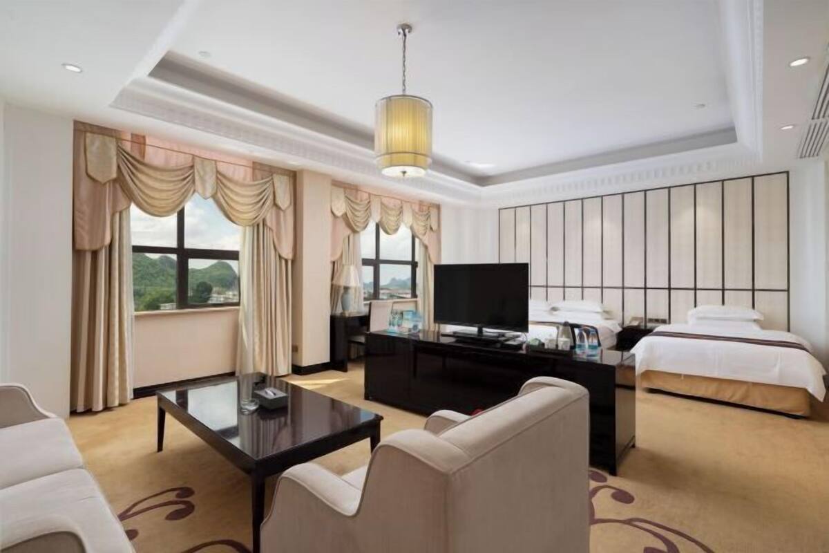 A hotel room with a bed, a TV, a desk, a sofa, and a large window with curtains.