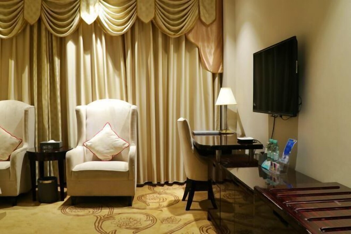 A hotel room with a sofa, armchair, desk, TV, lamp, and a carpeted floor.