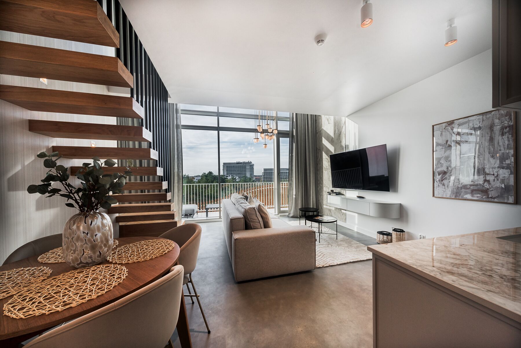 Een moderne woonkamer met een houten trap, een eethoek met een tafel en stoelen, een bank, een aan de muur bevestigde televisie en een groot raam met uitzicht op de stad.