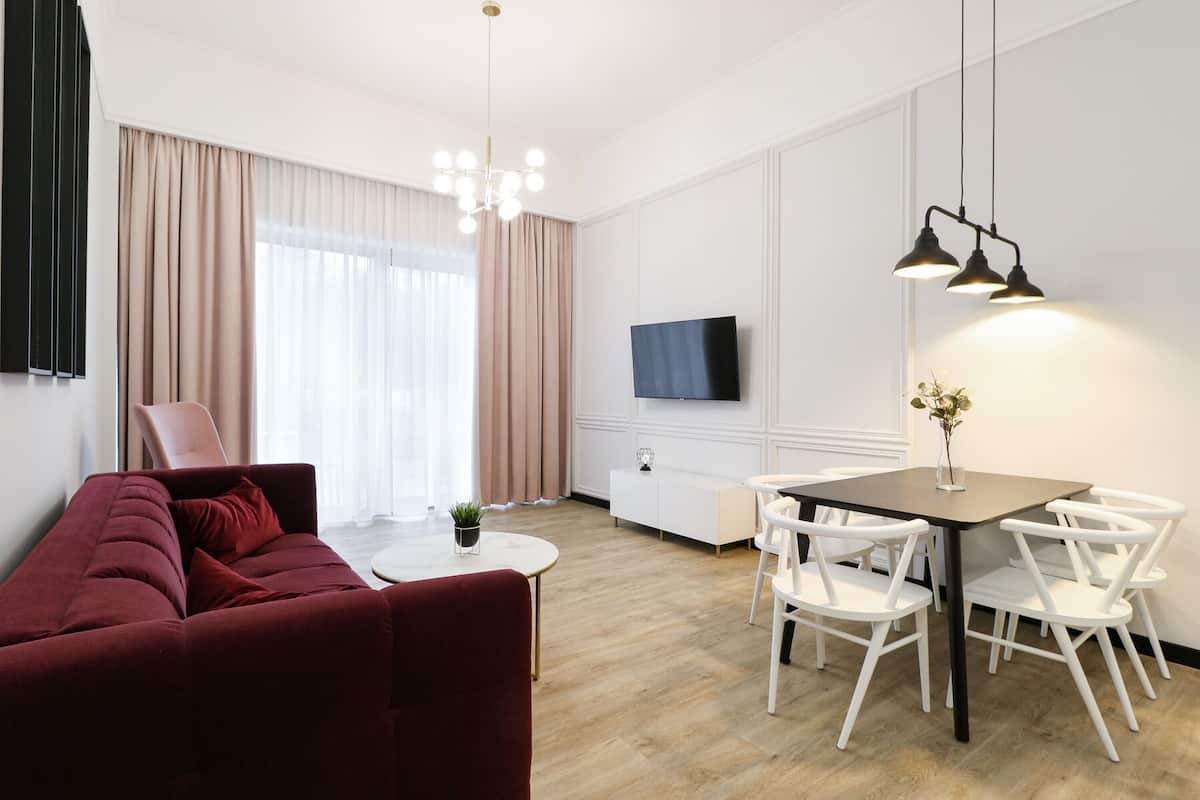 Ein modernes Wohnzimmer mit einem roten Sofa, einem weißen Esstisch mit weißen Stühlen, einem an der Wand befestigten Fernseher und einer Kristallleuchtre.