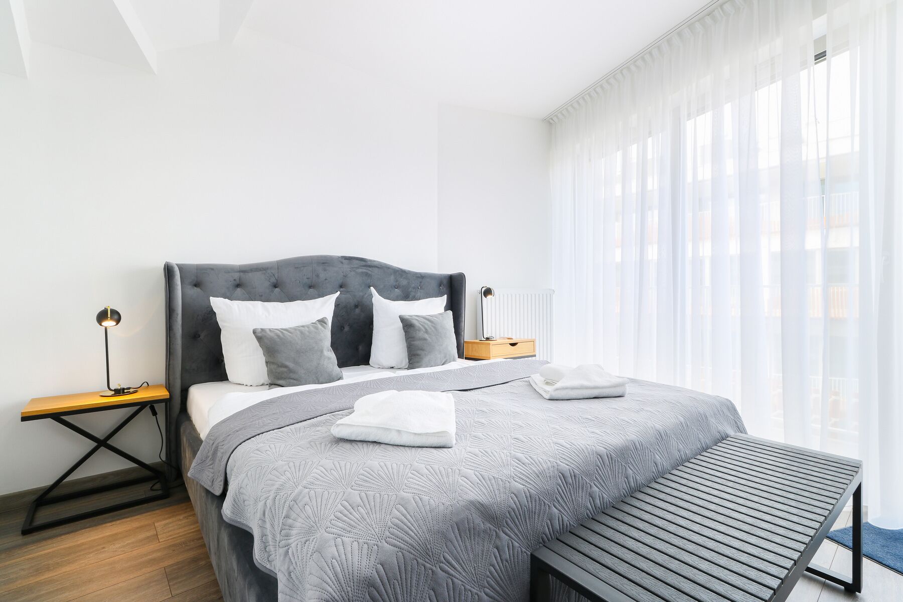 Un dormitorio moderno con cabecera capitonada, colcha estampada gris, mesita de noche con lámpara y un banco.