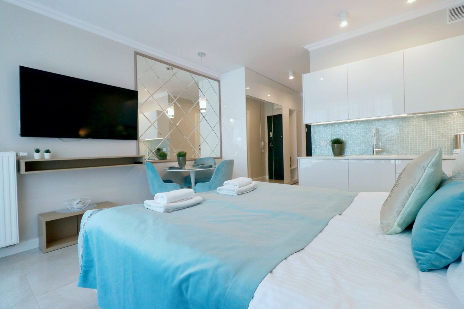 A modern hotel room with a large bed, a flat-screen TV mounted on the wall, a small dining table with chairs, and a kitchenette area.