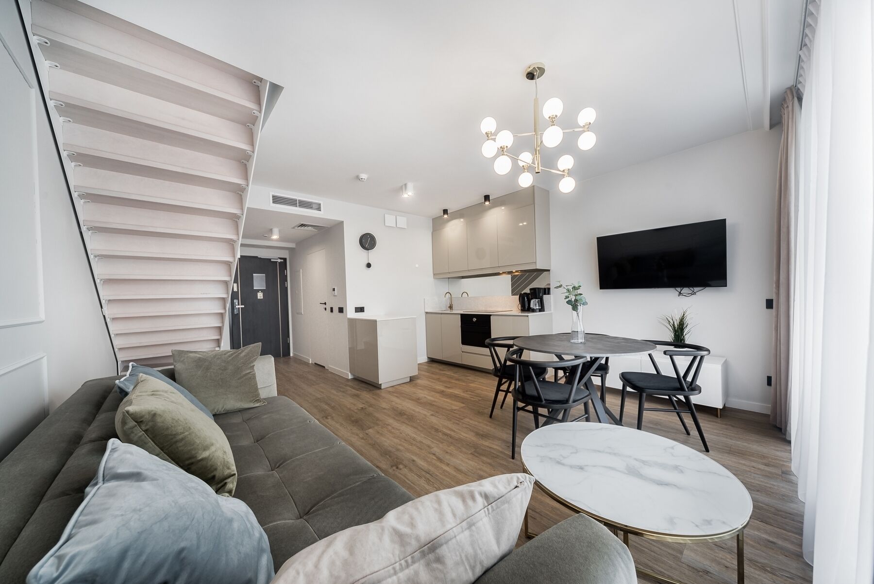 Een moderne woonkamer met een grijze bank, een ronde marmeren tafel en een keukenhoek met een televisie en een kroonluchter.