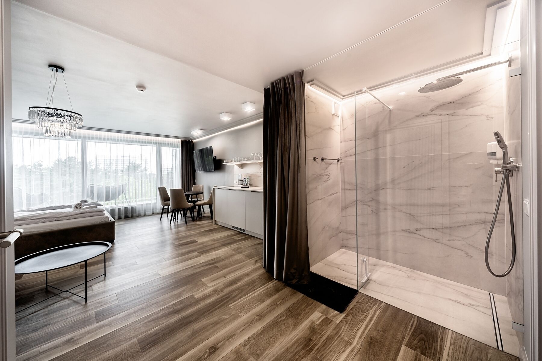 Et moderne hotelværelse med trægulv, et spiseområde med bord og stole, et fjernsyn ophængt på væggen og en bruseniche med glasdør.