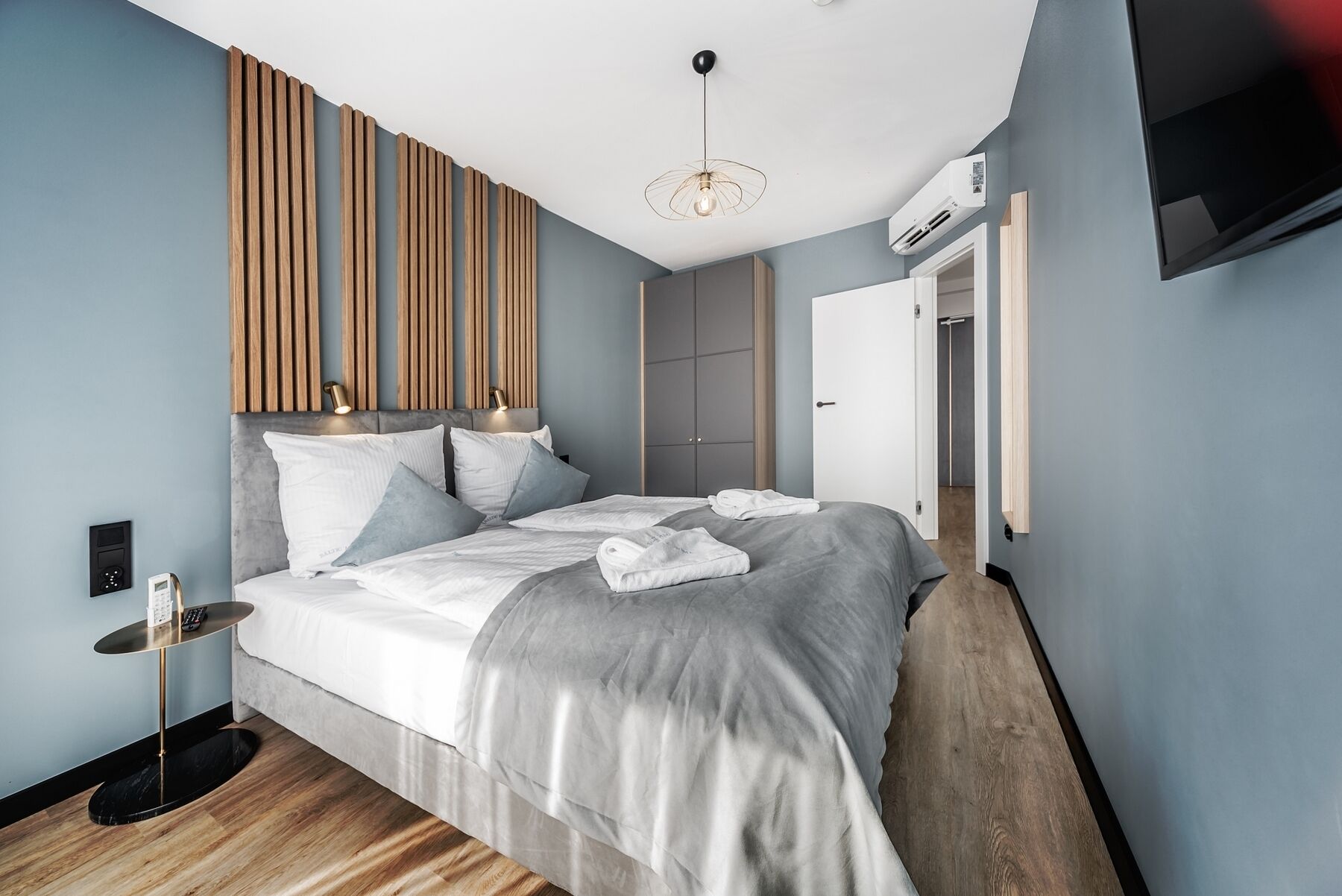 A modern bedroom with a bed, bedside tables, a wooden slat wall, a ceiling light, and an air conditioning unit.