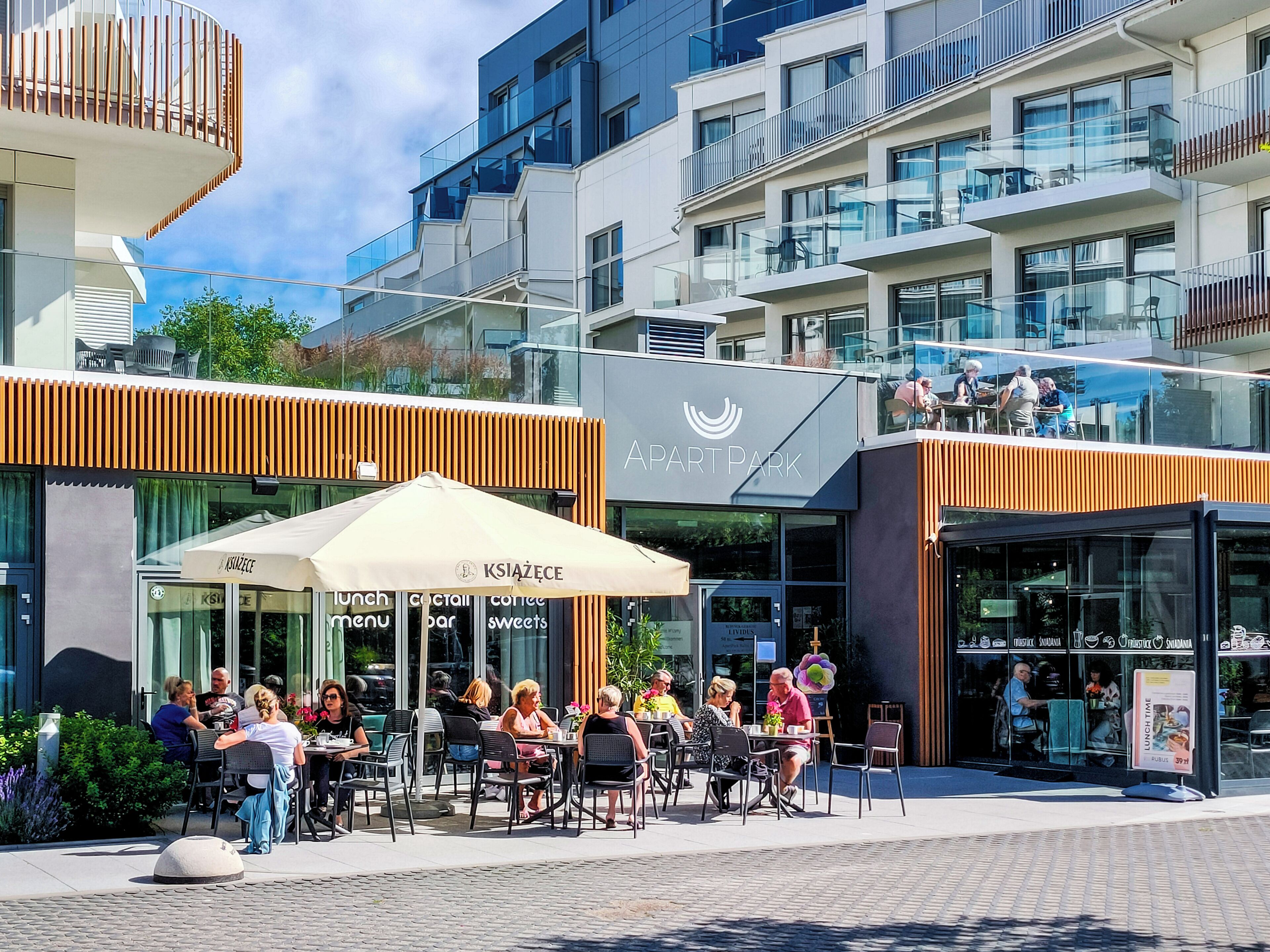 An outdoor cafe with tables and chairs, a sign for 'KSIAŻECE' offering lunch, cocktails, and sweets, and a building with balconies and glass railings in the background.