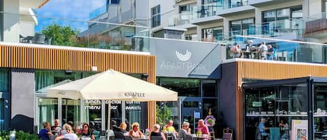 Un café al aire libre con mesas y sillas, un letrero que indica 'KSIAŻECE' ofreciendo almuerzos, cócteles y dulces, y un edificio con balcones y barandillas de cristal al fondo.