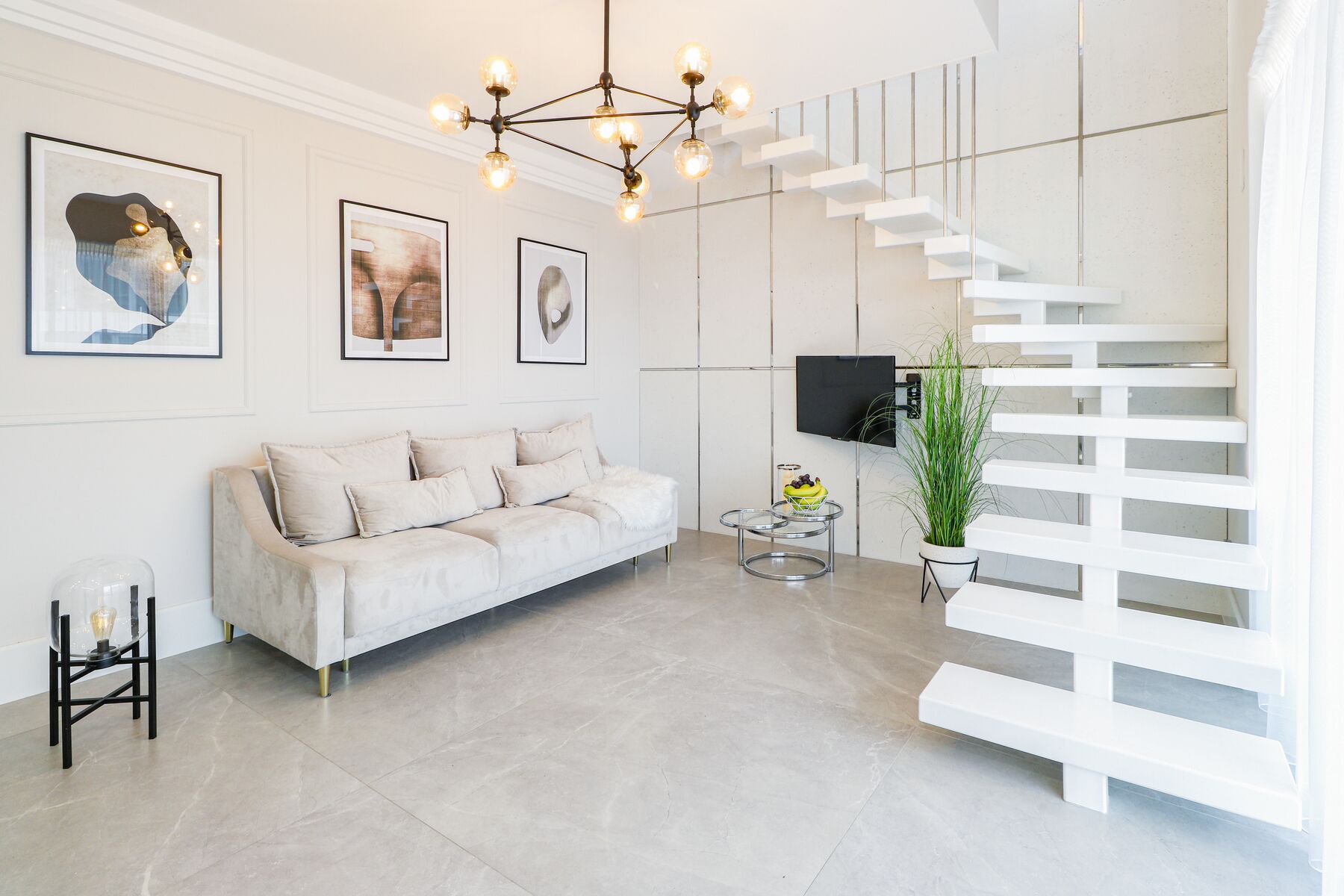 A modern living room with a white sofa, a glass coffee table, a chandelier, and a staircase.